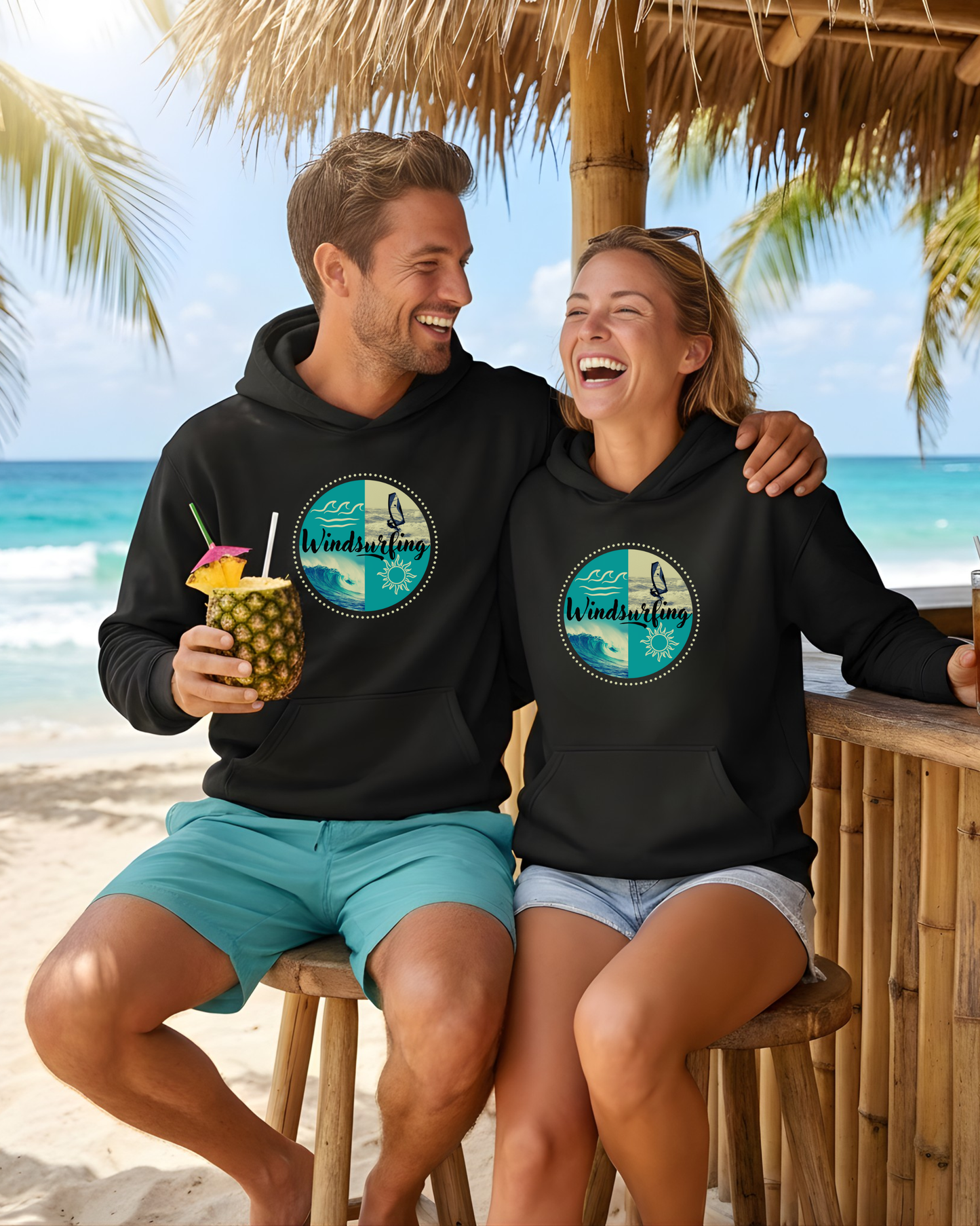 Ein junges Pärchen an einer Beachbar am Strand. Beide tragen einen schwarzen Bio-Hoodie mit einem Windsurfing-Design.
